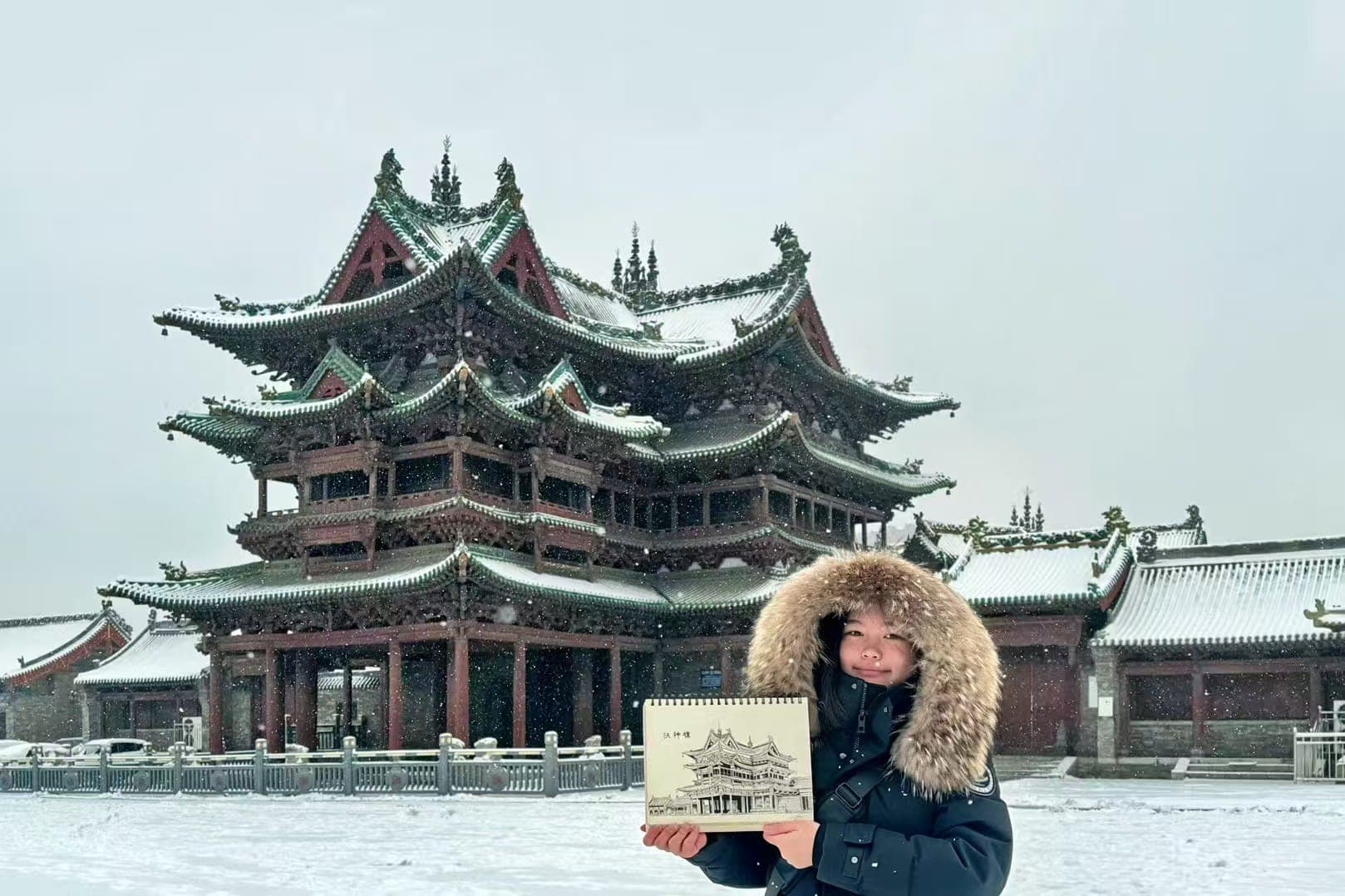 Gloria holding an architectural drawing in front of a snow-covered traditional Chinese building
